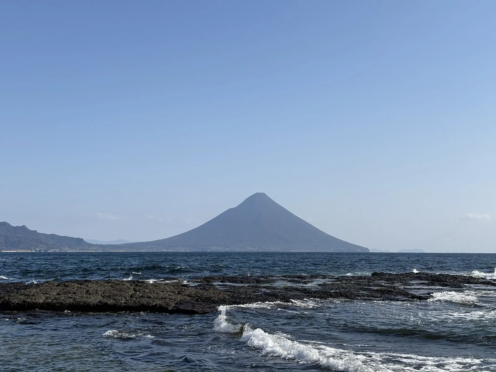 番所鼻の岩礁に寄せる波と、その向こうに開聞岳